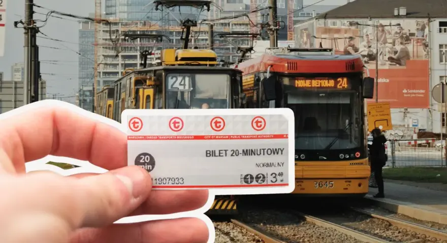 Warszawski bilet 20 minutowy w dłoni, na tle tramwajów i miejskiej zabudowy.
