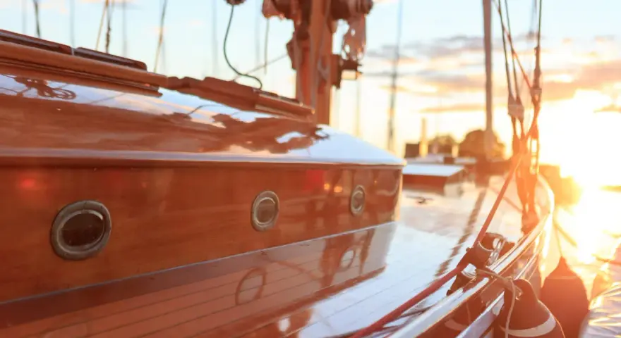 Glänzendes Holzdeck eines Segelboots, frisch lackiert. Kleine Bullaugen spiegeln die untergehende Sonne wider.