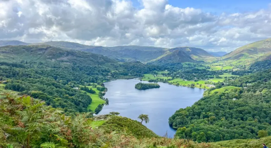 A stunning view of a lake surrounded by green hills and trees, perfect for day trips from Liverpool. A lone hiker walks down a path in the foreground.
