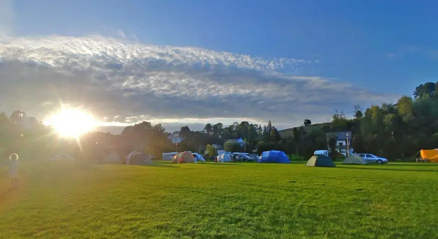 Słońce przebija się przez chmury nad polem namiotowym. Rozstawione namioty i samochody zapowiadają udany camping.