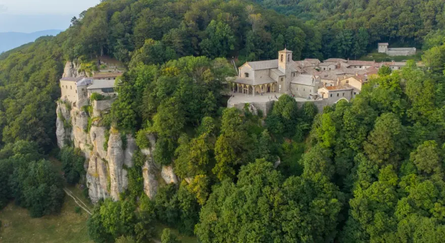 A monastery complex clings to a sheer cliff face in La Verna, Italy, surrounded by lush green forests.