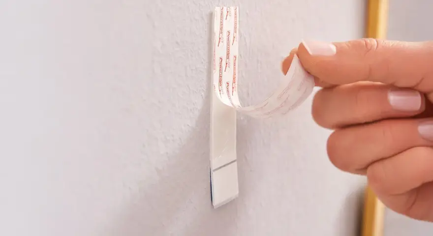 Dłoń odrywa taśmę z napisem "Powerstrips", pokazując alternatywę dla gwoździ do ściany.
