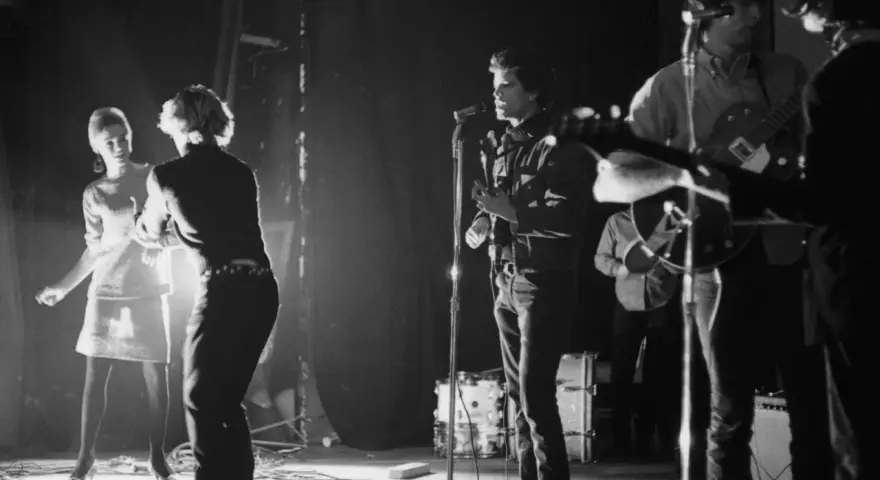 Scène de concert des membres de The Velvet Underground. Une femme danse à gauche, tandis que Lou Reed chante au micro.
