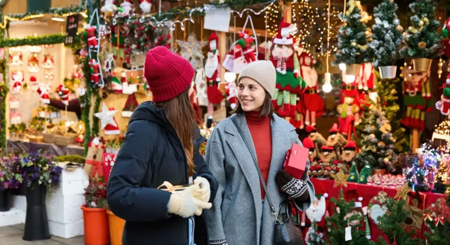 Två kvinnor njuter av julmarknader i Skåne, omgivna av julpynt och ljus. De håller presenter och ler mot varandra.