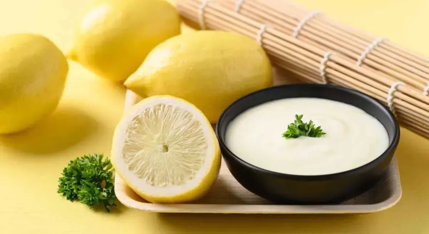 Un bol de sauce beurre citron crémeuse, garni de persil, avec des citrons frais et un rouleau de bambou en arrière-plan.