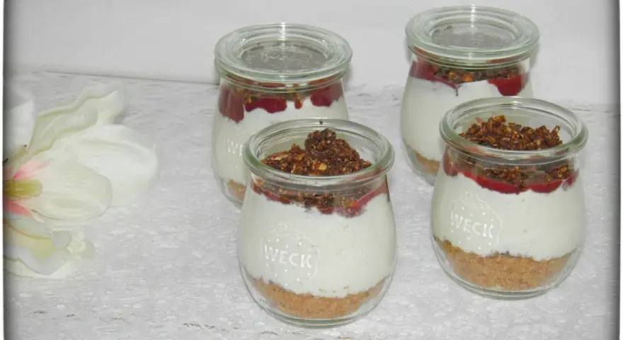 Vier leckere Desserts im Glas mit Krümelboden, cremiger Füllung, roter Fruchtsauce und knusprigem Topping.