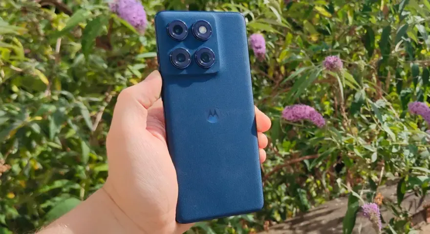En hand håller en mörkblå motorola edge 60 fusion med fyra kameror mot en bakgrund av gröna blad och lila blommor.