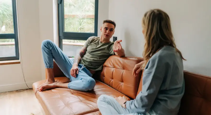 Un homme et une femme discutent sur un canapé en cuir marron. Leurs vêtements décontractés suggèrent un moment de détente, peut-être un double lien familial ou amical.