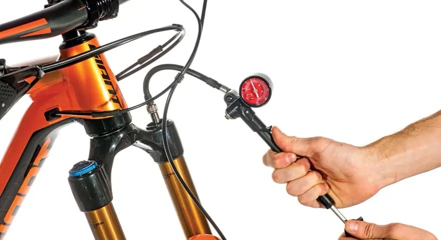 Adjusting the mtb suspension setup. A hand uses a shock pump with a gauge to set the air pressure on an orange mountain bike's front fork.