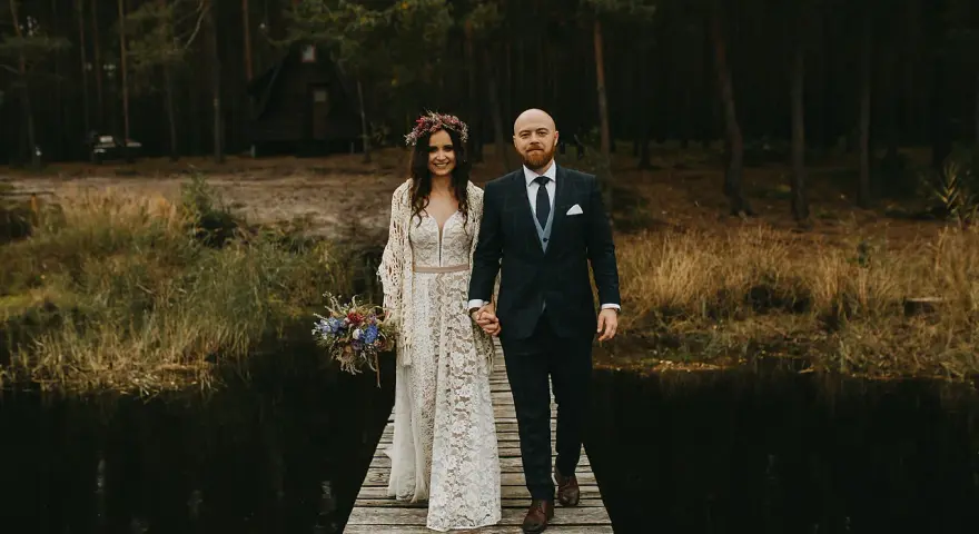 Para idzie przez las, ona w koronkowej sukni i wianku, on w garniturze. Ich suknia ślubna boho rustykalna idealnie pasuje do leśnej scenerii.