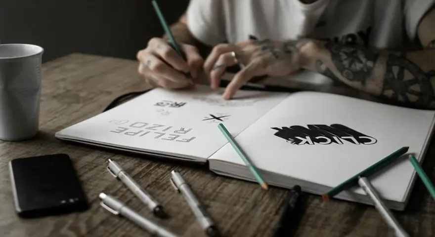 Un artiste auteur ou micro entrepreneur dessine dans son carnet, entouré de stylos, crayons et d'une tasse.