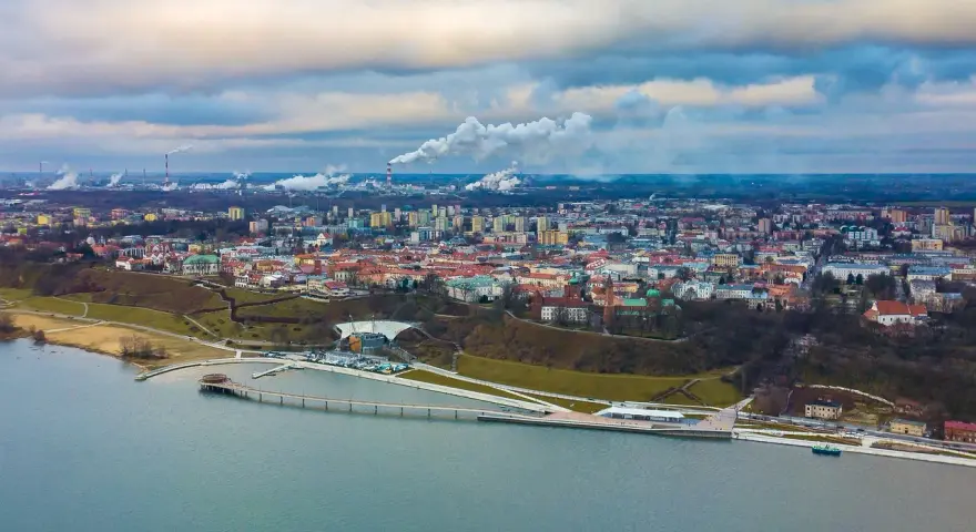 Widok na Płock z lotu ptaka, z Wisłą w tle. Widać panoramę miasta, fabryki z dymiącymi kominami i nowoczesną infrastrukturę nad rzeką, która może być częścią atrakcji dla dzieci w Płocku.