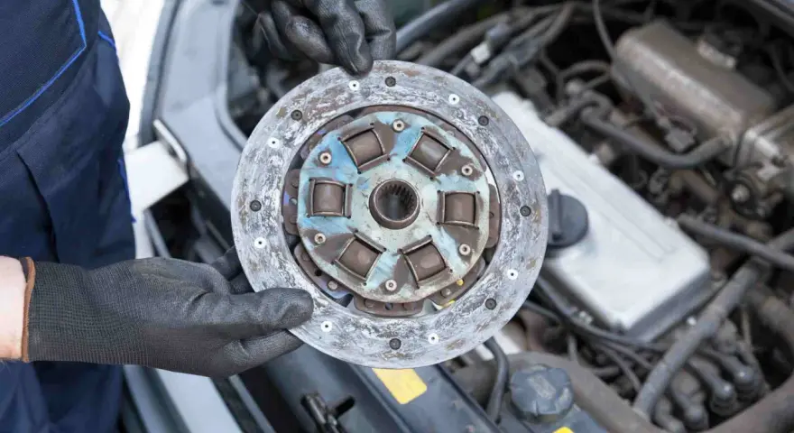 Mechanik w czarnych rękawiczkach trzyma zużyte sprzęgło samochodowe na tle silnika.