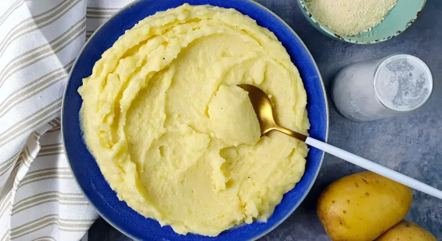 Un piatto di cremoso pure di patate, pronto per essere gustato. Accanto, patate fresche, sale e formaggio grattugiato.