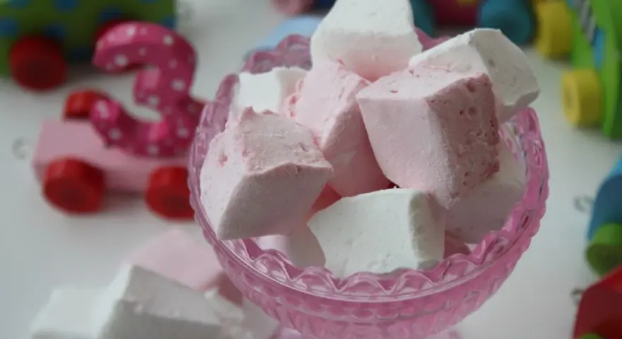 En skål med hemmagjorda marshmallows i rosa och vitt. I bakgrunden syns färgglada träleksaker, som en rosa bil med siffran 3.