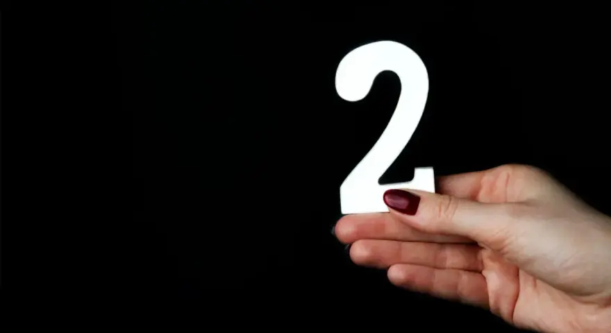 Dłoń z ciemnoczerwonym lakierem trzyma białą cyfrę 2. Co oznacza liczba 2?