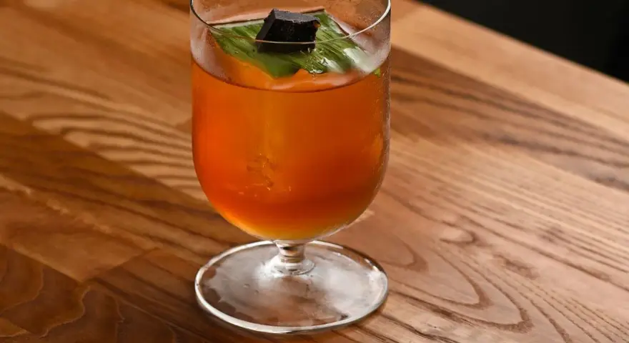 Klasyczny Mai Tai z sokami, ozdobiony liściem i kostką ciemnego rumu, na drewnianym blacie.