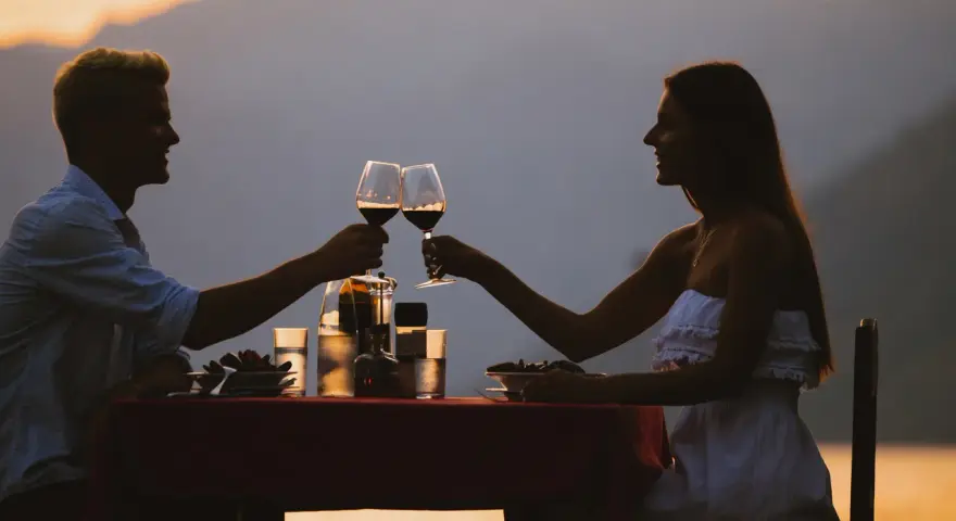 Idealne miejsce na pierwszą randkę: para wznosi toast winem przy romantycznej kolacji na tle zachodzącego słońca i gór.