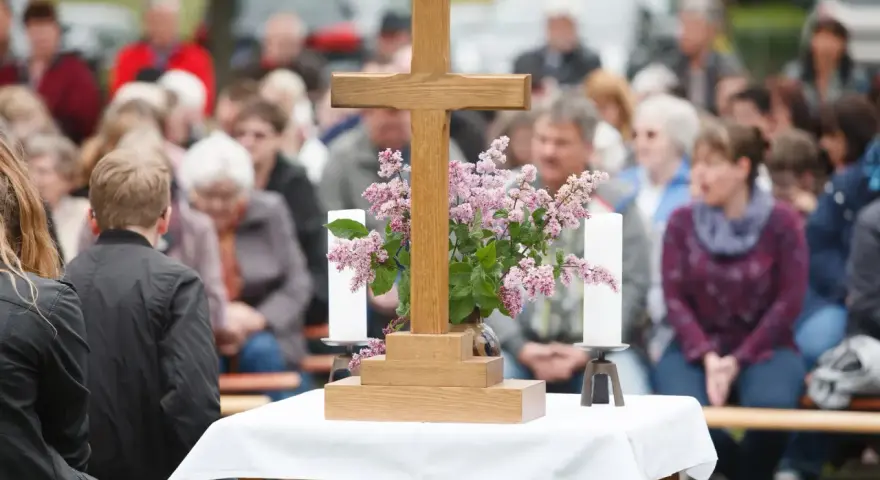 Holzkreuz mit Blumen und Kerzen vor einer Gemeinde. Ein Thema, das auch bei unterschiedlichen Konfessionen von Ehepartnern relevant ist: die Kirchensteuer.