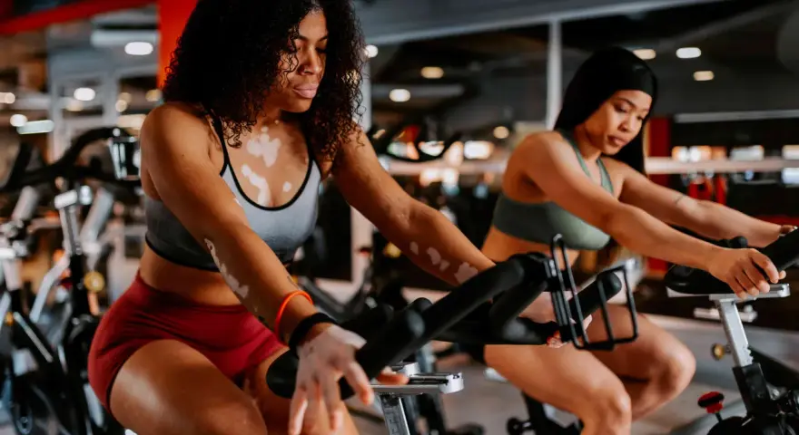 Two women are engaged in a stationary bike workout in a gym. The woman in the foreground has vitiligo and curly hair.