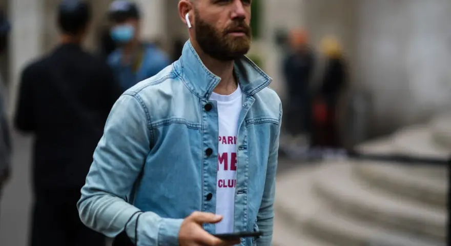 Hombre calvo sin barba, con auriculares y chaqueta vaquera, mira su móvil.