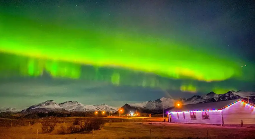 Islandia pogoda listopad: zorza polarna nad ośnieżonymi górami i domkiem z lampkami.