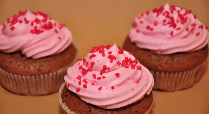 Tre chokladmuffins med rosa frosting och röda strössel, bakade i rosa muffinsformar.