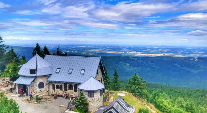 Kamienny schronisko z szarym dachem w Górach Izerskich, otoczone zielonymi lasami i rozległą panoramą dolin pod błękitnym, chmurowym niebem.