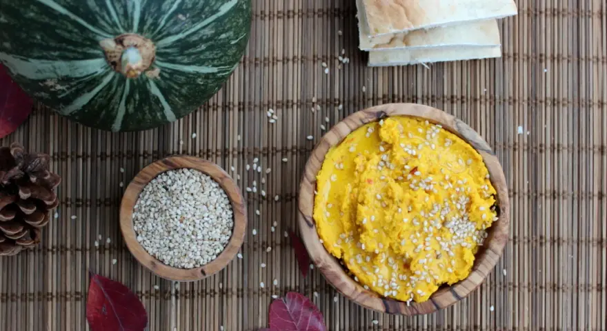 Ein leuchtend gelber Hummus, bestreut mit Sesam, wartet neben einer Schale voller Sesam und einem Kürbis. Perfekt für ein kleines Hummus-Baby-Festmahl!