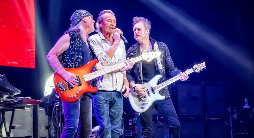 Trzech muzyków na scenie w głębokim fioletowym świetle Atlas Areny. Jeden śpiewa, dwaj grają na gitarach.