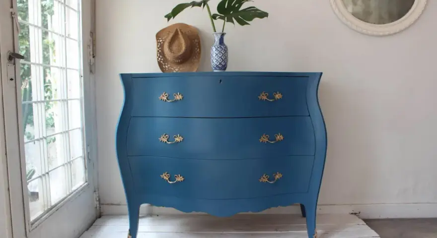 Cómoda antigua con **muebles pintados en azul** y tiradores dorados, decorada con un sombrero y un jarrón con hojas.