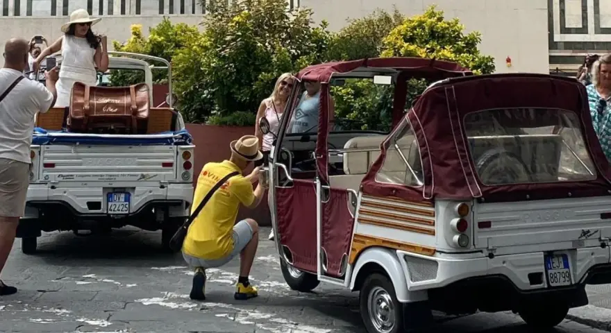 Turisti scattano foto a veicoli atipici, un Piaggio Ape e un tuk-tuk, in una strada soleggiata.