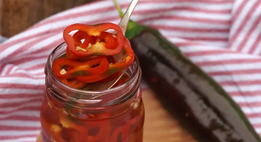 En burk med picklad chili, med en sked som lyfter upp en skiva. En mörk chili ligger bredvid.