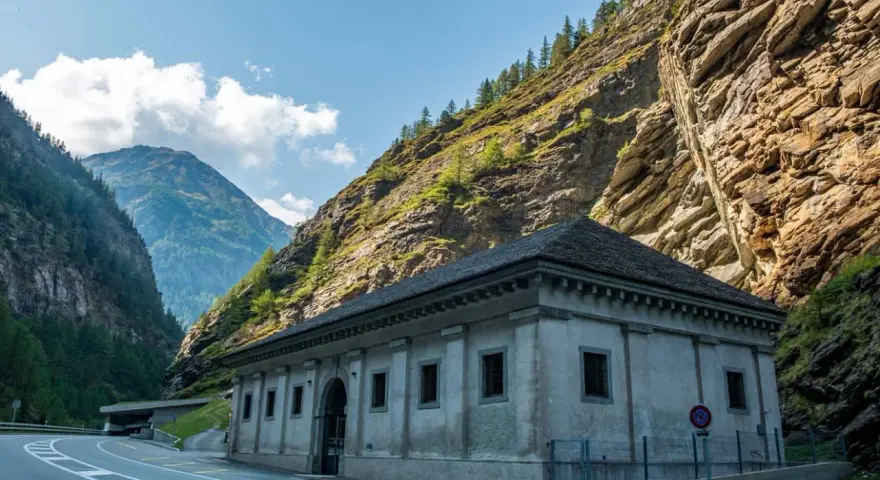 Edificio in pietra ai piedi di una montagna rocciosa, con strada e tunnel. Un passo alpino che sembra non finire mai, un vero sempione passo.
