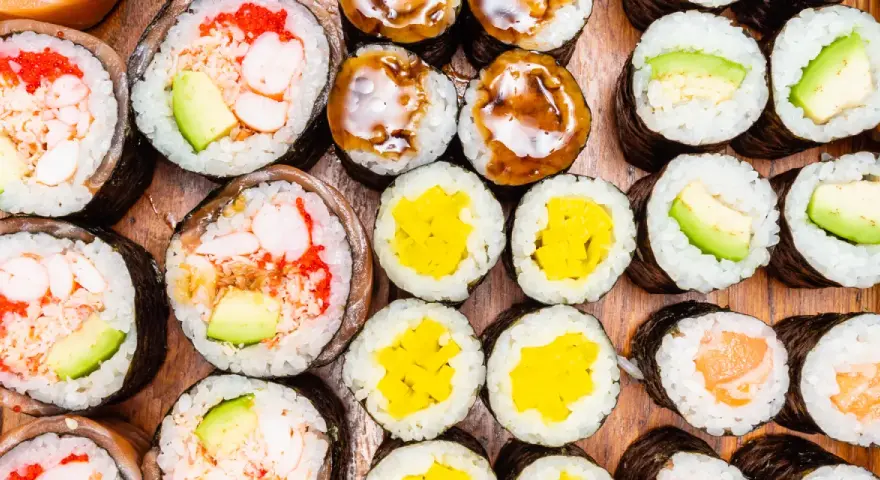 Różnorodne rolki sushi: grubsze futomaki z krewetkami i awokado, cieńsze hosomaki z łososiem, awokado, żółtą rzodkwią i polane sosem.