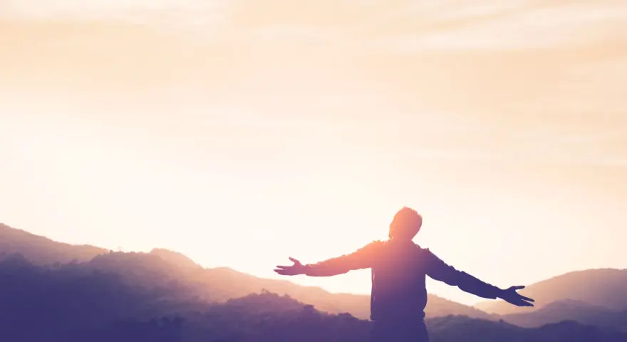 Silhouette einer Person mit ausgebreiteten Armen vor einer Bergkulisse bei Sonnenuntergang. Ein Symbol für Kraft und Hoffnung nach einer Krankheit.