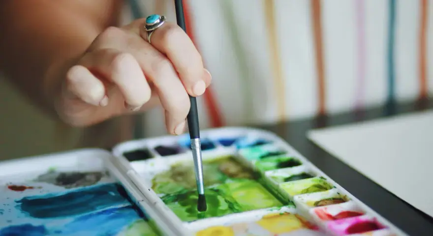 Main d'une artiste, bague turquoise, tenant un pinceau au-dessus d'une palette de couleurs vives de **de l'aquarelle**.