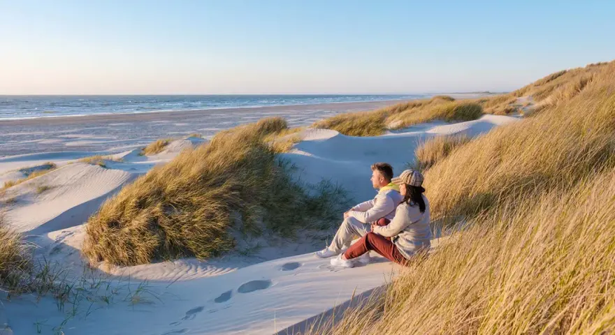 Ett par sitter på en sanddyn och njuter av utsikten över havet. Vackra sanddyner med gräs sträcker sig längs kusten, en idyllisk plats i en nationalpark i Danmark.