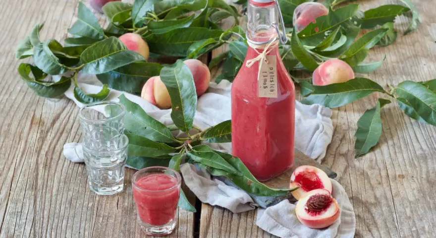 Flasche mit selbstgemachtem Pfirsichschnaps, frische Pfirsiche und Blätter auf Holztisch. Perfekt für den Sommer!