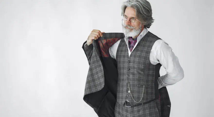 Elegancki mężczyzna z siwą brodą i wąsami, ubrany w kraciasty garnitur, prezentuje się jak prawdziwy arbiter elegantiarum.