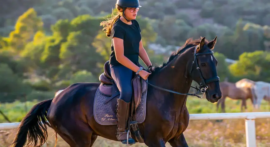 Jeune fille en casque et lunettes roses monte un cheval noir dans un ranch, le soleil couchant illumine la scène.