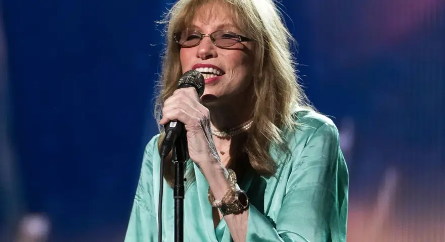 Carly Simon heute, mit Brille und Perlenkette, singt ins Mikrofon. Ihr seidiges, türkisfarbenes Kleid fällt elegant.
