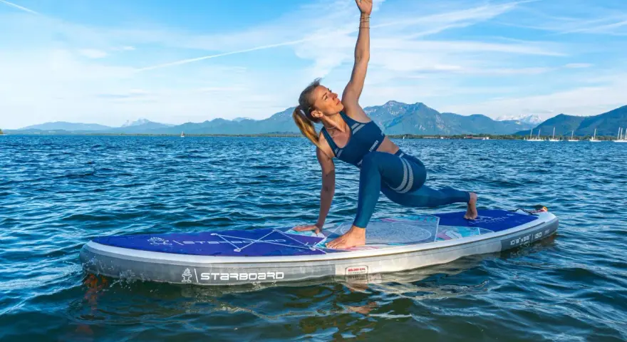Frau praktiziert Krieger II auf einem SUP-Board auf ruhigem Wasser, Berge im Hintergrund.