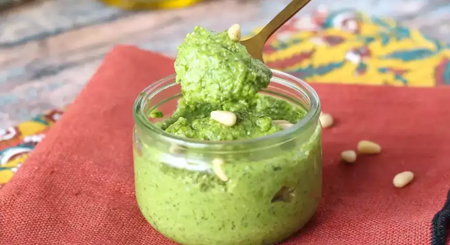 Łyżka nabiera aromatyczne pesto z rukoli z małego słoiczka. Na czerwonym tle widać orzeszki piniowe.