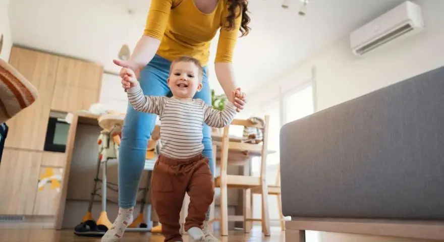 Mamá ayuda a su bebé a dar sus primeros pasos en una habitación cómoda, con el aire acondicionado bebes funcionando para mantener la temperatura ideal.
