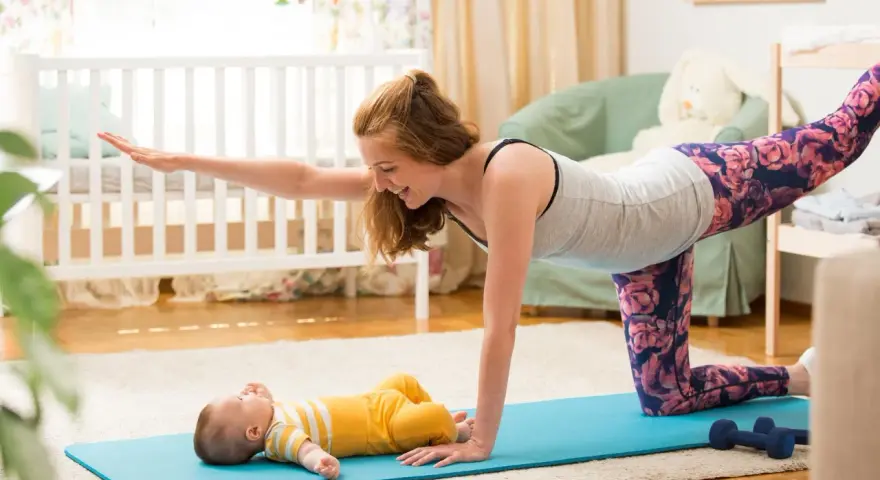 Mama wykonuje ćwiczenia po porodzie na macie, a jej maluch leży obok.