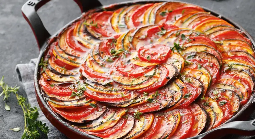 Ein wunderschönes Ratatouille-Rezept, das in einer roten Pfanne mit geschichteten Tomaten-, Zucchini- und Auberginenscheiben und frischen Kräutern präsentiert wird.