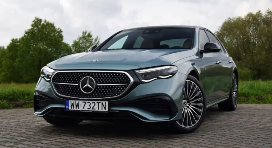Elegancki Mercedes E220d w kolorze miętowym, zaparkowany na brukowanej nawierzchni, z zielenią w tle.