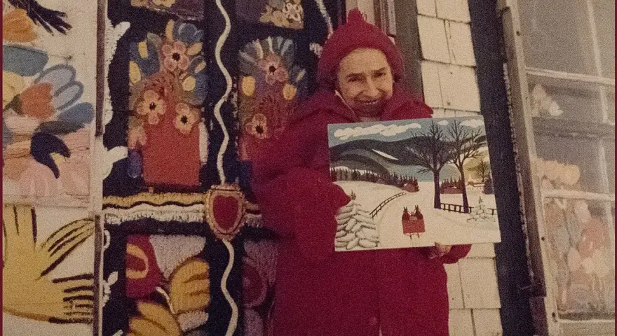 Maud Lewis, klädd i rött, håller upp en av sina charmiga vintermålningar framför sitt färgstarka hem.