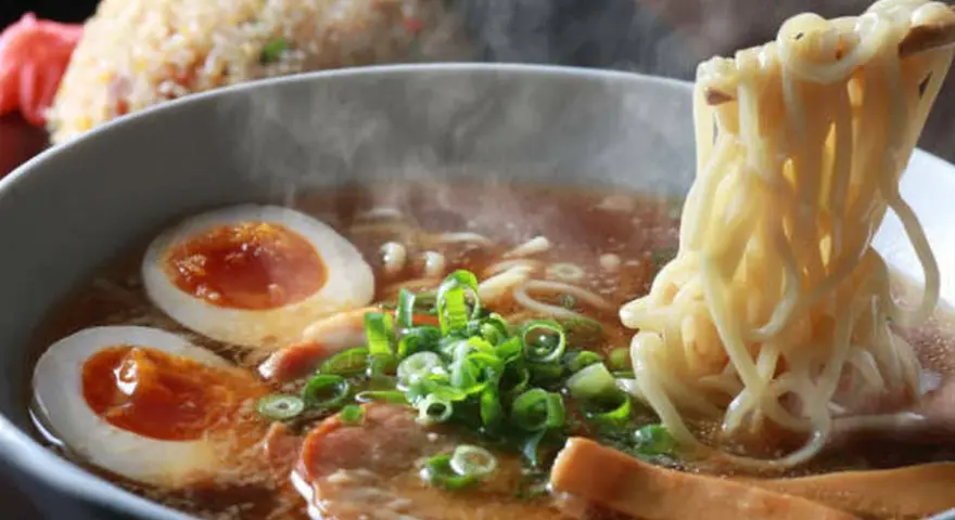 Un humeante tazón de ramen con huevo cocido, cebolleta y cerdo. Los palillos levantan fideos, listos para disfrutar de esta deliciosa receta ramen.
